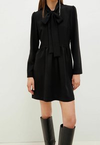 Frau in einem schwarzen Langarm-Minikleid mit einer großen Schleifendetail am Hals und schwarzen kniehohen Stiefeln, die vor einem schlichten Hintergrund steht.