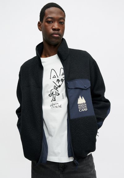 Μαλακή ζακέτα από φλις σε μαύρο χρώμα με ναυτικό τσέπη. Διαθέτει φερμουάρ, υψηλό γιακά και λεπτομέρεια με λογότυπο. Συνδυάζεται με λευκό μπλουζάκι με γραφικά.