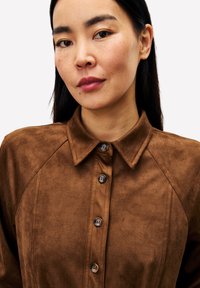 Frau mit schmalem schwarzen Haar, die ein braunes, knöpfbares Wildledershirt trägt und mit einem neutralen Gesichtsausdruck leicht zur Kamera schaut.