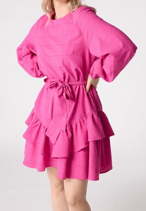 Mujer con un vestido de manga larga de capas color rosa brillante, con cinta en la cintura, de pie con las manos en las caderas contra un fondo liso.