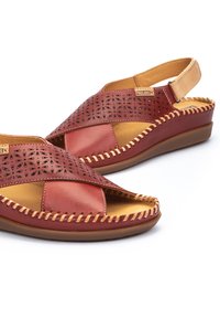 Sandalias de cuero rojo con un diseño recortado, que cuentan con un interior marrón y un acento trenzado alrededor de la suela. Correa ajustable en el talón.