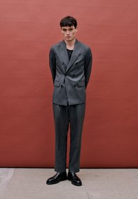 Halli kahekordne ülikond, struktureeritud õlgadega, kohandatud püksid ja mustad nahasandaalid. Taust on ühtlane punane.