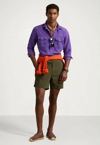 Μωβ πουκάμισο, πορτοκαλί πουλόβερ δεμένο στη μέση, πράσινα σορτς, εσπαντρίγιες. Αξεσουάρ περιλαμβάνουν γυαλιά ηλίου και μια με patterned μαντήλι.