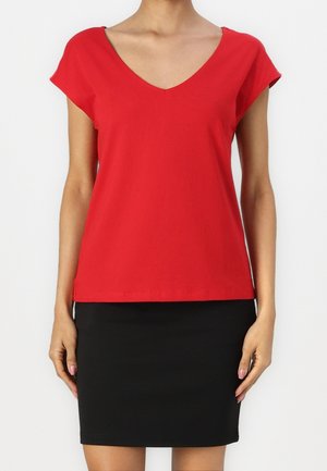 Femme portant un haut rouge à manches courtes avec col en V et une jupe noire longueur genou, debout sur un fond blanc.