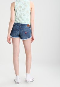 Persona che indossa una canotta leggera con stampa di palme verde chiaro, pantaloni shorts di denim sfilacciati con un patch sul retro e sneakers bianche alte, in piedi di spalle.