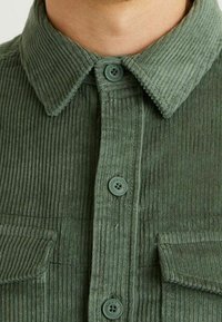 Vihreä corduroy-paita, jossa on napillinen kaulus, kaksi rintataskua ja teksturoitu ribbineulos sekä vastaavat napit.