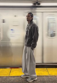 Harmaa corduroytakki, jossa etutaskut, väljät harmaat collegehousut ja harmaat tennarit, taustalla sumea metroympäristö.