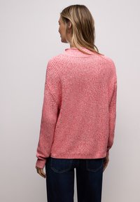 Femme portant un pull en maille rose et un jean bleu foncé, vue de dos sur un fond uni clair.
