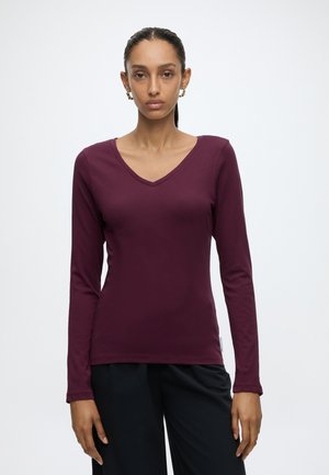 Femme debout devant un fond uni, portant un haut bordeaux à manches longues avec un col en V et un pantalon noir, face à l'appareil avec une expression neutre.