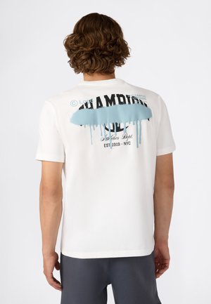 Persoon met krullend bruin haar die een witte t-shirt met korte mouwen draagt, voorzien van een blauwe verfspetter en zwarte tekst op de achterkant.