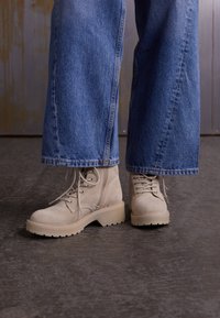 Beige ruskind lace-up støvler med kraftige såler, parret med vide, blå denim jeans. Støvlerne har en struktureret overflade og fremtrædende synsømme.