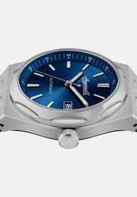 Automatyczny zegarek ze niebieską tarczą, srebrną kopertą i oknem daty. Posiada srebrne znaczniki godzin, prostokątne indeksy oraz teksturowaną koronkę.