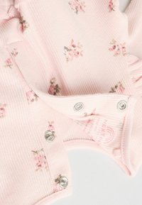 Rosa gerippter Stoff-Overall mit kleinen Blumenmustern, Druckknöpfen und einer weichen Textur. Markentag in der Nähe des Ausschnitts sichtbar.