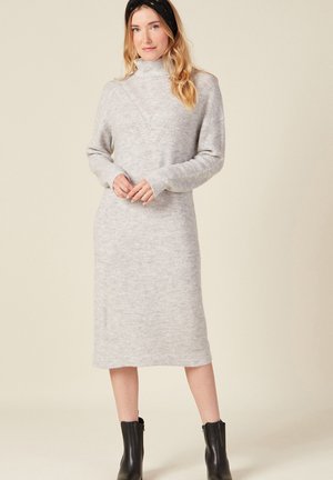 Grau gestreiftes Strickkleid mit hohem Kragen, langen Ärmeln und einem knielangen Saum. Schwarze Stiefeletten runden das Outfit ab.