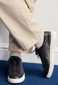 Črni usnjeni supergi z belo gumijasto podplatom, okroglo konico, vezalkami in teksturiranimi poudarki, kombinirani z bež hlačami in svetlimi nogavicami.