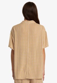 Chemise à manches courtes en tissu jaune doré avec un motif floral, dotée d'un col et d'une coupe décontractée, avec des détails de couture visibles au dos.
