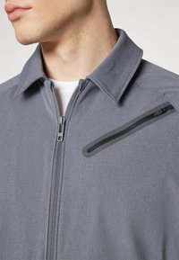 Gray zip-up jacket with a textured fabric, fold-down collar, and a zippered chest pocket featuring black trim and hardware accents.