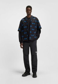 Kortearm zwart shirt met blauw logo-patroon, gelaagd over een zwart langemouwen top, gecombineerd met donkere cargobroek en zwarte sneakers.