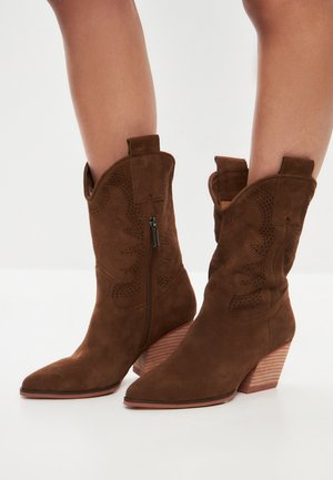 Las botas de cowboy de ante marrón presentan una punta afilada, costuras decorativas, un cierre lateral de cremallera y un tacón apilado con textura similar a la madera.