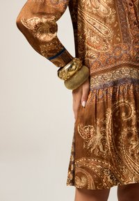 Bruin jurk met paisleypatroon, lange mouwen en een geplooide rok. Gouden armbanden sieren de pols en tonen ingewikkelde ontwerpen en texturen.