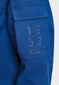 Manche de veste bleue avec une poche ornée des chiffres brodés "1 8 / 5 3" et du mot "AIGLE" en dessous.