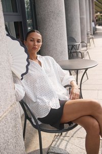 Blouse blanche à boutons perforés superposée sur une jupe noire. Modèle assis avec un éventail blanc à bord festonné et une table ronde à proximité.