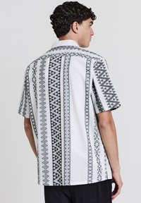Uomo con camicia bianca a maniche corte con strisce verticali geometriche nere, in piedi con la schiena rivolta verso la macchina fotografica su uno sfondo semplice.