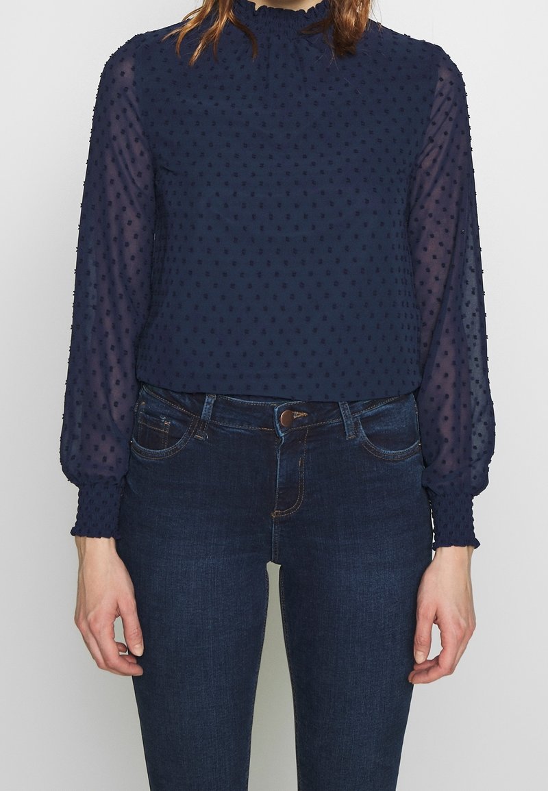 Navyblauwe blouse met een gestructureerd stippenpatroon, hoge nek en doorschijnende pofmouwen, gecombineerd met donkere spijkerbroeken.