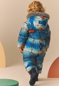 Mono de nieve acolchado azul con patrones geométricos multicolores, capucha forrada de pelo gris, cinturilla elástica y botas azul marino con suelas texturizadas.