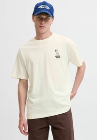 Cremefarvet bomulds T-shirt med korte ærmer, prydet med et sort grafisk print på venstre bryst. Båret med brune bukser og en blå baseballkasket.