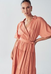 Persikkavärinen kietaisumekko, jossa on kaulus, lyhyet hihat ja vyötärölle sidottu vyö. Mekossa on kerroksellinen hameosa ja pehmeä, teksturoitu kangas.