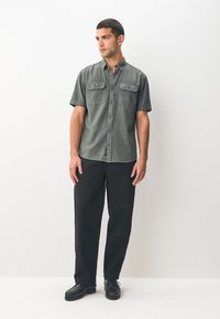Short-sleeve, dark green shirt with two chest pockets, button-up front, and a textured fabric. Paired with black, loose-fitting pants and black shoes.
