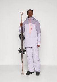 Žmogus lavandinės spalvos slidinėjimo striukėje ir kelniuose, laikantis slidžių porą vertikaliai, dėvintis juodus slidinėjimo batus, stovintis prieš paprastą foną.