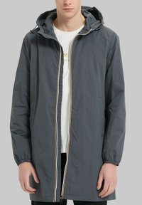 Veste imperméable grise avec capuche, tissu léger, poignets élastiques, fermeture éclair sur le devant avec une bande contrastante, et poches latérales.