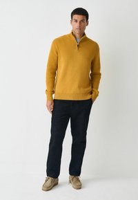 Senfgelber Reißverschluss-Pullover mit gerippten Bündchen und Saum, kombiniert mit schwarzen Cordhosen und beigen Bootsschuhen. Glatte Textur, einfaches Design.