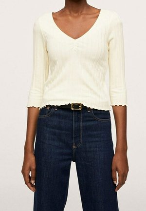 Frau trägt ein cremefarbenes, geripptes V-Ausschnittshirt mit 3/4-Ärmeln und dunkelblaue, hoch taillierte Jeans mit einem schwarzen Gürtel und goldener Schnalle.