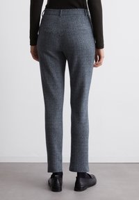 Un pantalon herringbone gris sur mesure présente une coupe slim, une texture lisse, une taille standard et deux poches arrière, associé à des chaussures noires.