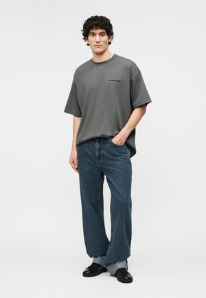 Jeune homme debout, une main dans la poche, portant un t-shirt gris ample avec un petit texte, un jean large bleu et des chaussures noires.