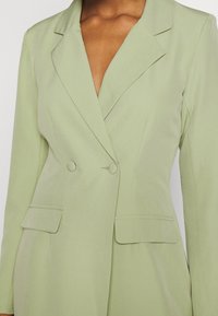 Ljusgrön kavaj med en figurvänlig design, med slagkrage, dubbla knappar och två framfickor. Slät textur och skräddarsydd form.