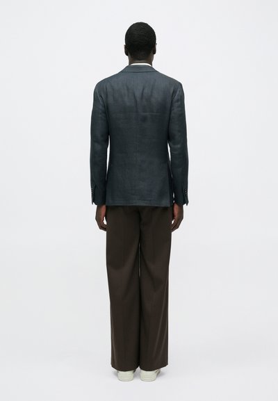 Άνδρας που στέκεται με την πλάτη γυρισμένη, φορώντας σκουρό γκρι σακάκι, καφέ φαρδιά παντελόνια και λευκά αθλητικά παπούτσια, μπροστά σε λευκό, απλό φόντο.