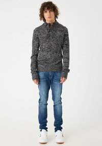 Suéter de punto gris oscuro para hombres con un cuello alto, que presenta un patrón texturizado y mangas largas, combinado con jeans azul de corte slim y zapatillas blancas.