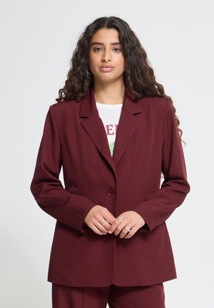 Donna con capelli lunghi e ricci che indossa giacca e pantaloni color bordeaux, in piedi davanti a uno sfondo bianco semplice.
