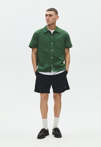 Jeune homme debout, les mains dans les poches, portant une chemise verte à manches courtes boutonnée, un short noir, des chaussettes blanches et des chaussures noires sur fond blanc.