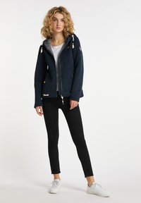 Marinfärgad fleecejacka med dragkedja fram, grått innerfoder, gula detaljer och justerbar huva. Matchad med svarta jeans och vita sneakers.