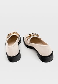 Béžové boty z umělé kůže s kulatou špičkou, černou gumovou podrážkou a zlatým řetízkem přes vršek.