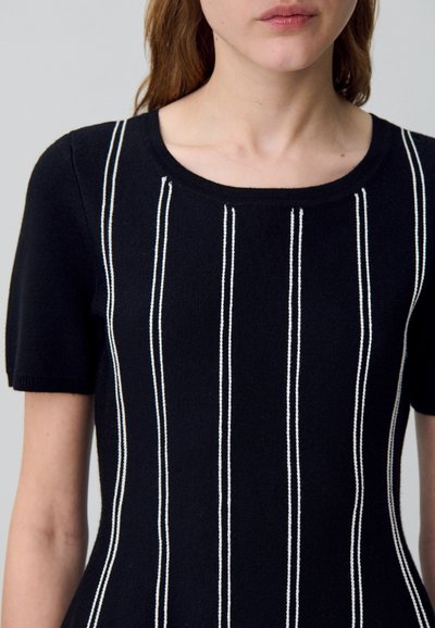 Mujer que lleva una blusa negra de manga corta con rayas blancas verticales, escote redondo y silueta ajustada, mostrada desde el cuello hasta la cintura.