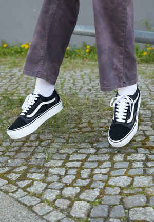 Baskets en suède noir avec des accents blancs, présentant un bout rond, une semelle blanche épaisse et un design classique à lacets, portées avec des chaussettes blanches et un pantalon violet.