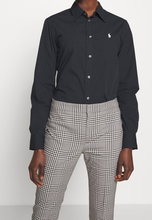 Persona con una camisa negra abotonada con un logo blanco y pantalones de cuadros en blanco y negro de tiro alto, sobre un fondo liso.