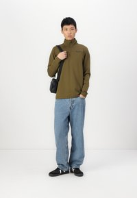 Langärmliger, olivgrüner Pullover mit einem Halbzip-Kragen. Getragen mit hellblauen, weit geschnittenen Jeans und schwarzen Schuhen. Kleine schwarze Schultertasche.