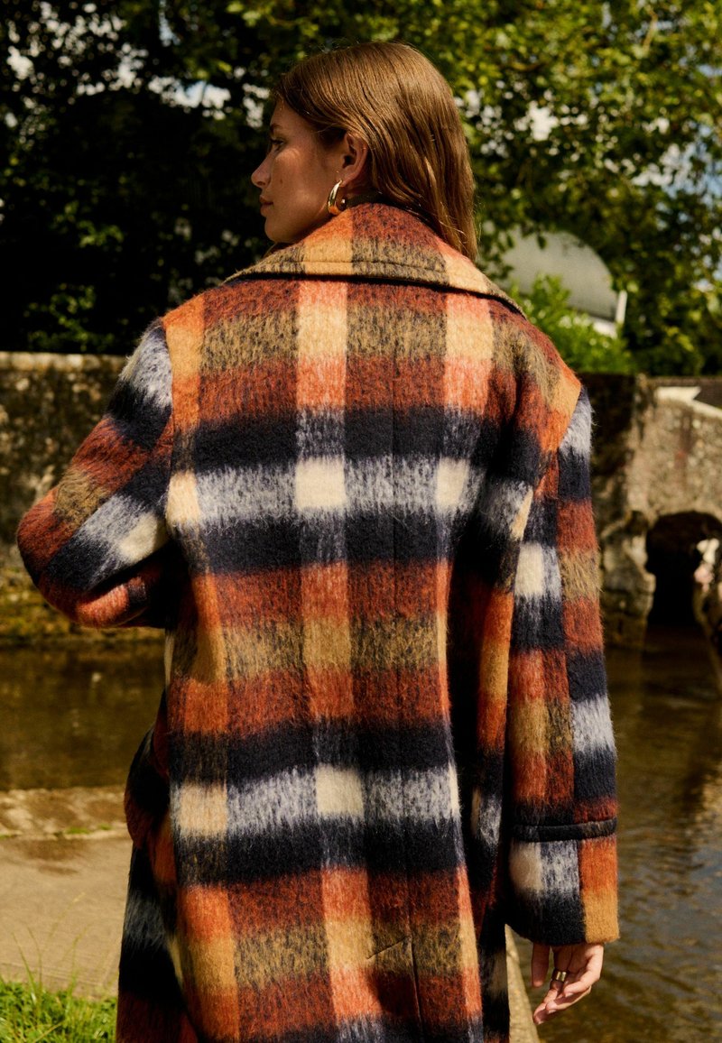 Manteau en laine à carreaux avec des couleurs orange, marine et beige, doté d'un col, de longues manches et d'une coupe décontractée. Tissu texturé avec de grands motifs à carreaux.
